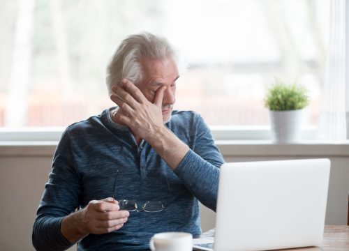 Fatigued mature old man taking off glasses suffering from tired dry irritated eyes after long computer use, senior middle aged male feels eye strain problem or blurry vision working on laptop at home