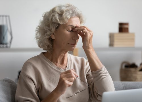 Exhausted senior older woman taking off glasses, suffering from dry eyes syndrome after computer overwork, sitting alone on sofa in living room. Middle aged grandmother rubbing eye, feeling tired.
