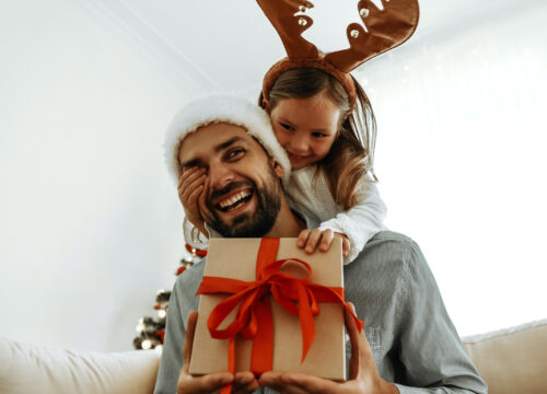Photo of a man opening a Christmas present with his daughter hanging on his shoulders